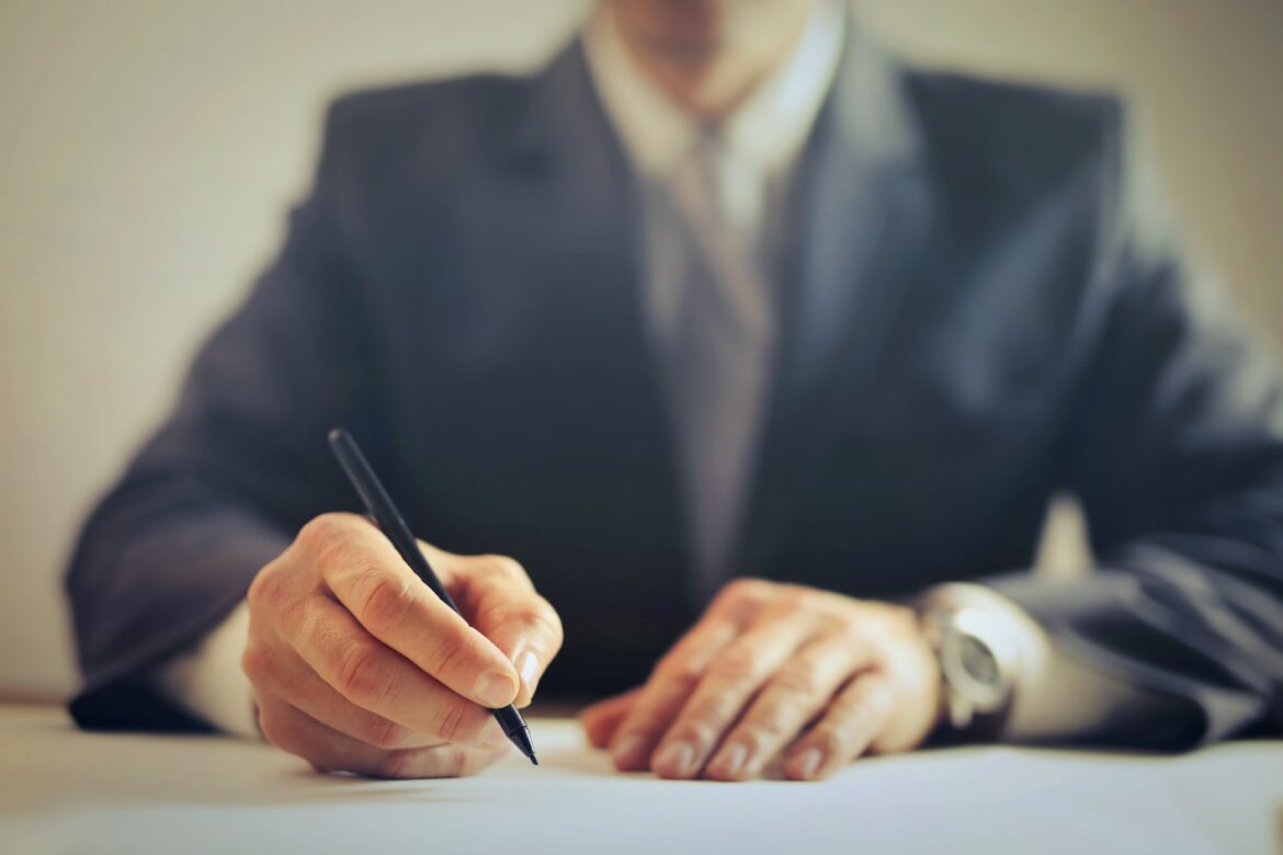 venditore senza volto in giacca che firma un atto di comprevendita sulla scrivania di un notaio